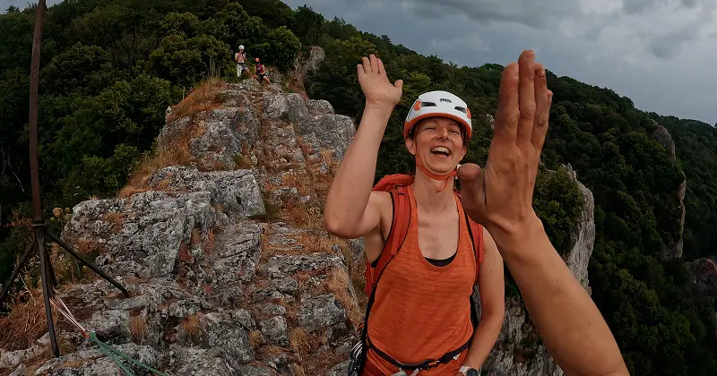 Een glimlachende bergbeklimmer in een oranje shirt en met een helm geeft een high-five op een rotsachtige bergkam, met weelderig groene bomen en andere bergbeklimmers op de achtergrond onder een bewolkte hemel.